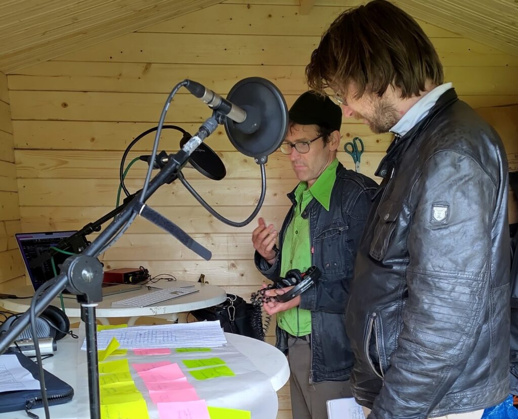 Zwei Männer stehen vor einem Tisch mit Mikro und Papieren auf dem Tisch.