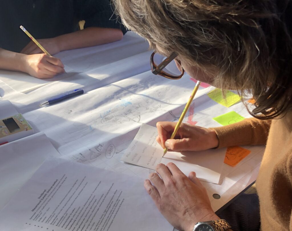 Blick über die Schulter einer Frau. Sie schreibt mit Bleistift auf einen Zettel. Weitere Papiere liegen auf dem Tisch.