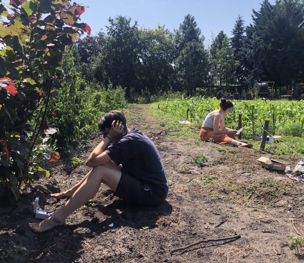 Ein Mann und eine Frau sitzen in etwas Entfernung voneinander auf dem Erdboden am Rand von Feldern. Der Mann hält Kopfhörer an seine Ohren. Die Frau schreibt etwas.